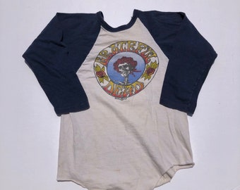 May include: Cream-colored baseball style shirt with navy blue sleeves. The front features a circular graphic with the words "Grateful Dead" surrounding a skull with roses. A vintage, retro style tee.