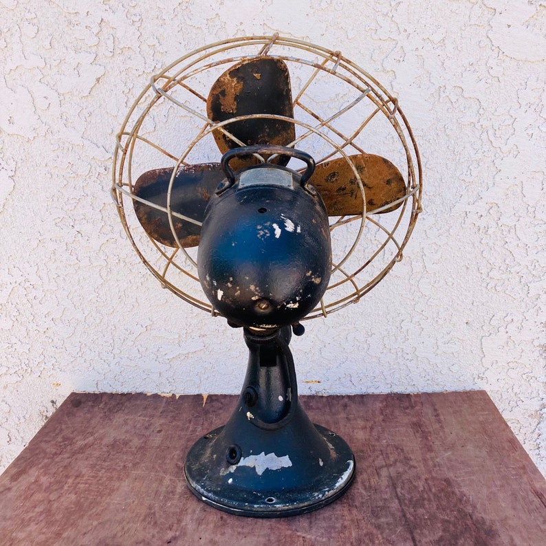 Pu&ograve; includere: Un ventilatore vintage in metallo nero con una gabbia di filo arrugginito e tre pale. Il ventilatore &egrave; su una superficie di legno.