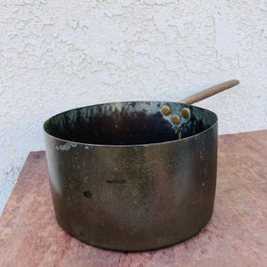May include: A vintage copper pot with a wooden handle. The pot has a dark patina and is slightly dented. It has three brass rivets on the side.