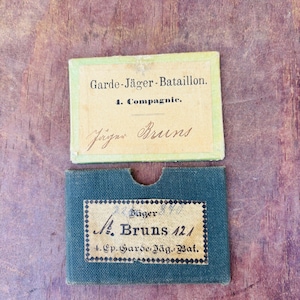 May include: Two antique military documents. The top document is a light green rectangle with the text "Garde-Jäger-Bataillon. 1. Compagnie. Jäger Bruns." The bottom document is a dark blue rectangle with the text "Jäger A. Bruns 121 4. Ep. Garde Jäg. Bat."