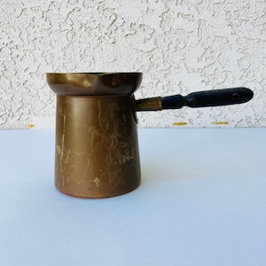 Puede incluir: Una cafetera de cobre vintage con mango de madera. La cafetera tiene un aspecto desgastado y envejecido, ideal para preparar café turco.