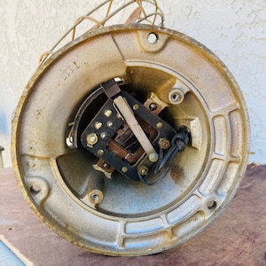 Pu&ograve; includere: Primo piano di un motore di ventilatore vintage in metallo con fili a vista e una gabbia in metallo. Il motore &egrave; dipinto di un colore oro chiaro e ha una forma circolare. Il motore &egrave; montato su una superficie di legno.