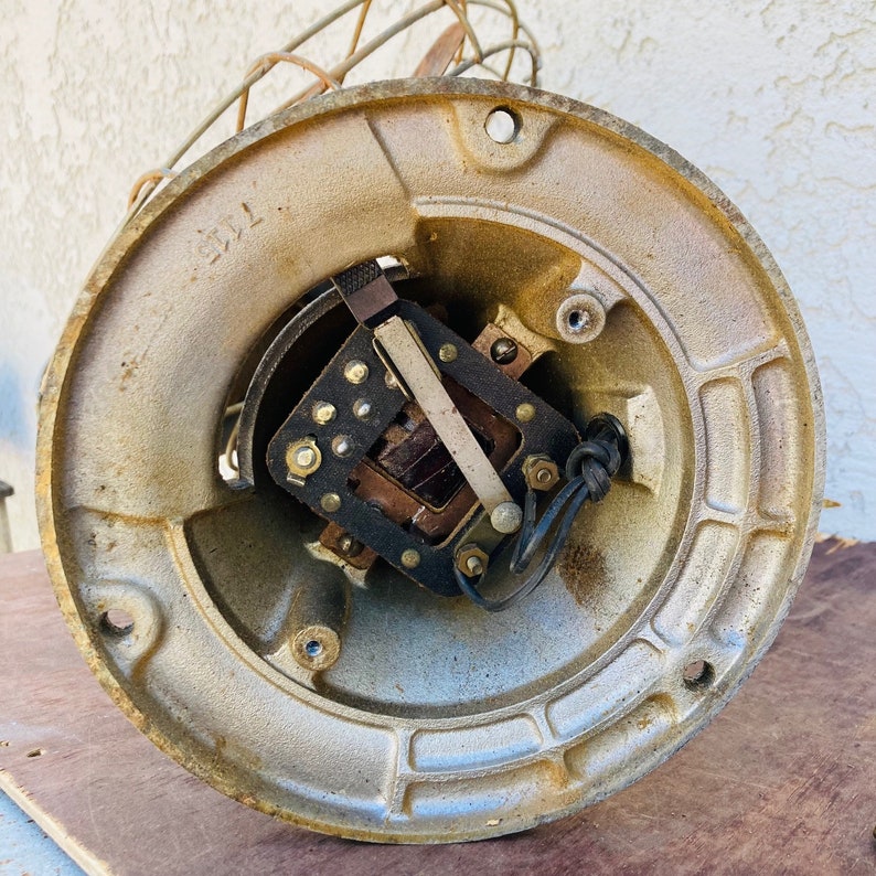 Pu&ograve; includere: Primo piano di un motore di ventilatore vintage in metallo con fili a vista e una gabbia in metallo. Il motore &egrave; dipinto di un colore oro chiaro e ha una forma circolare. Il motore &egrave; montato su una superficie di legno.