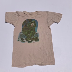 May include: A beige t-shirt with a graphic print of a green jester holding a guitar. The jester has a skull face and is wearing a green hat. The graphic is set against a blue and white checkered background.
