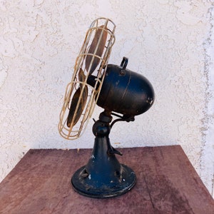 Pu&ograve; includere: Un ventilatore vintage in metallo nero con una gabbia di filo metallico e pale in legno. Il ventilatore &egrave; su un tavolo con una superficie in legno.