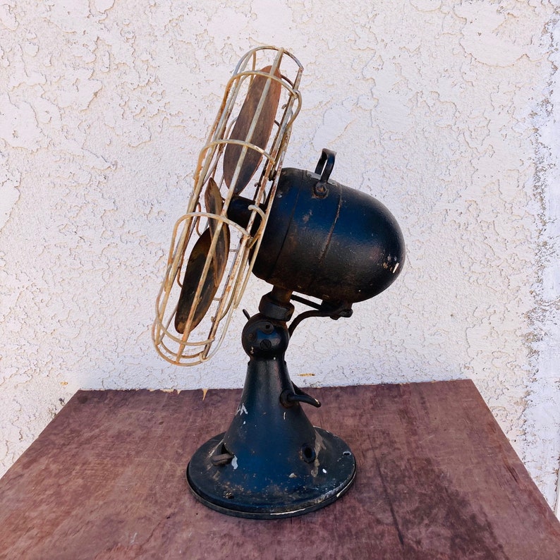 Pu&ograve; includere: Un ventilatore vintage in metallo nero con una gabbia di filo metallico e pale in legno. Il ventilatore &egrave; su un tavolo con una superficie in legno.