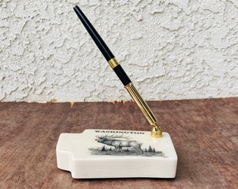 Könnte beinhalten: Ein weißer Keramik-Stiftständer mit goldfarbener Basis und einem schwarzen Stift. Der Ständer zeigt eine Zeichnung eines Elchs und das Wort "WASHINGTON".