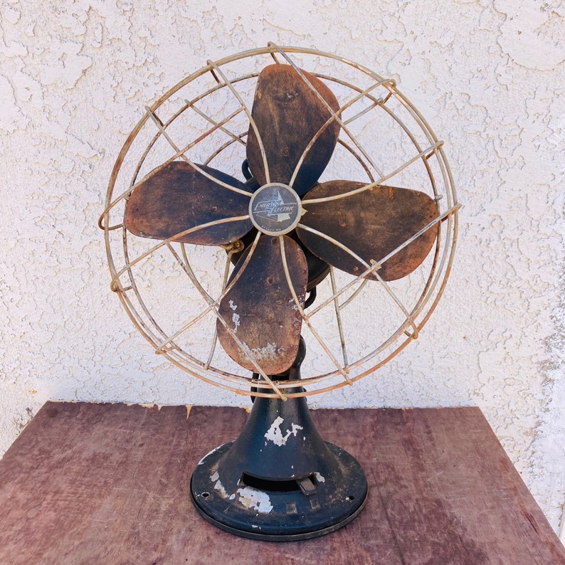 Pu&ograve; includere: Un ventilatore vintage in metallo con quattro pale nere e una gabbia argentata. Il ventilatore ha una base nera e un'etichetta argentata che recita "Emerson Electric Co."