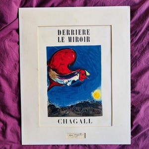 Pode incluir: Uma impressão emoldurada com uma obra de arte vibrante com uma paleta de cores vermelha, azul e amarela. A impressão inclui o texto "DERRIERE LE MIROIR" e "CHAGALL". A obra de arte retrata uma cena surrealista com figuras e uma paisagem urbana.