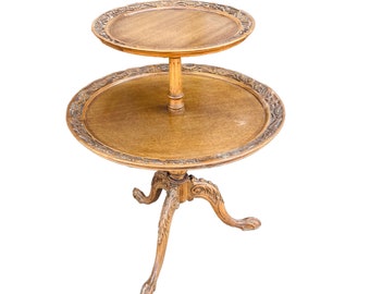May include: A two-tiered wooden table with a carved design on the top and bottom tiers. The table has three legs with carved feet.