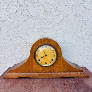 May include: A vintage wooden mantel clock with a light brown finish. The clock face is round with black numerals and hands, set within a dark metal frame. The clock's wooden body has a curved top and a rectangular base.