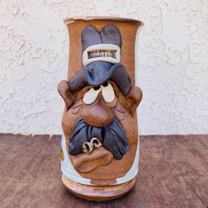 May include: A brown ceramic mug with a cartoon face, mustache, and hat. The hat has the word "WASTED" on it.