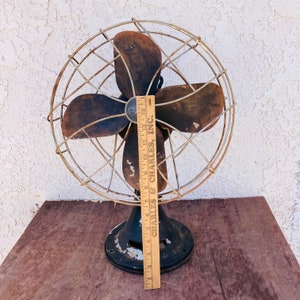 Pu&ograve; includere: Ventilatore vintage in metallo nero con tre pale marroni e una gabbia di filo. Il ventilatore &egrave; su una superficie di legno e ha un righello accanto con il testo "Charles & Charles, Inc."