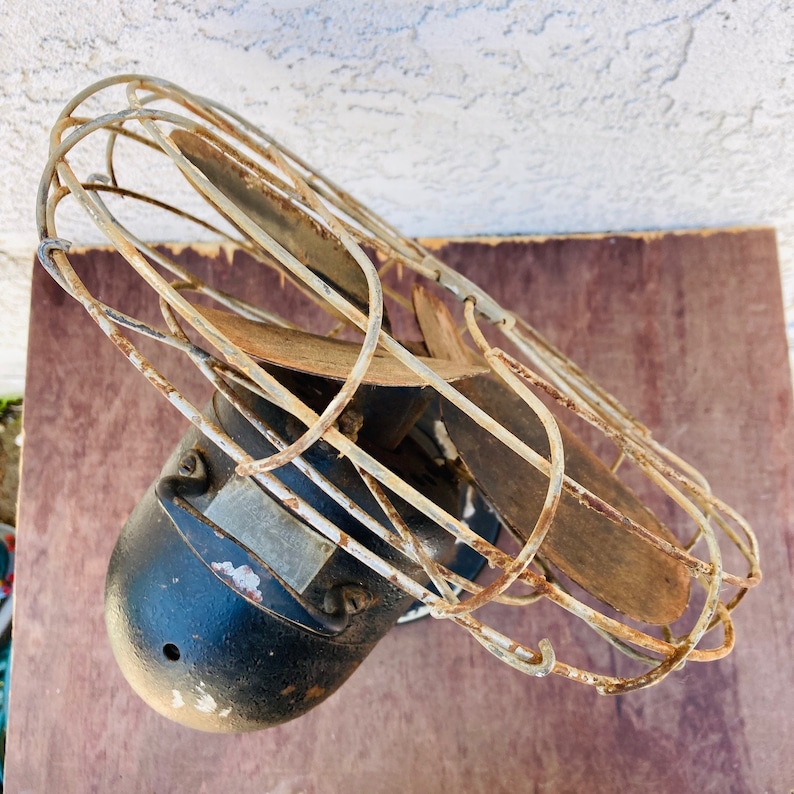 Pu&ograve; includere: Un ventilatore vintage in metallo nero con una gabbia di filo arrugginito e tre pale. Il ventilatore &egrave; montato su una parete e ha uno sfondo in legno marrone.