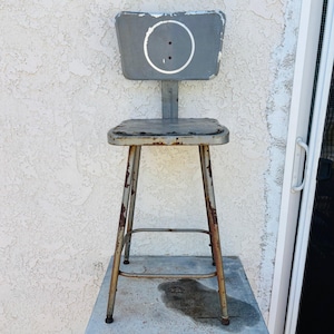 May include: Vintage industrial metal stool with a gray seat and backrest. The backrest features a white circle with two dots. The legs are metal with rust-colored patches. The stool is designed for seating.