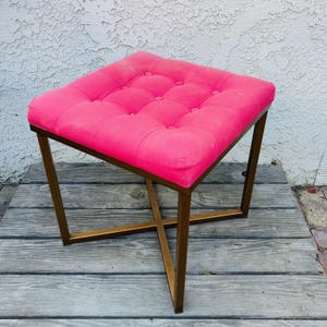 May include: A pink tufted ottoman with a gold metal frame. The ottoman has a square top and a cross-shaped base.