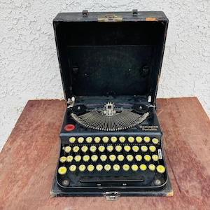 May include: Black Remington Portable typewriter with yellow keys, open in its black carrying case. The typewriter sits on a wooden surface. The text "Remington Portable" is visible on the typewriter.