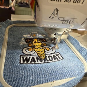 May include: A white embroidery machine stitching a yellow and grey mammoth graphic with the text WANKDAT onto a piece of blue denim fabric held in a rectangular embroidery hoop. The machine features a sticker with the text But so do I.