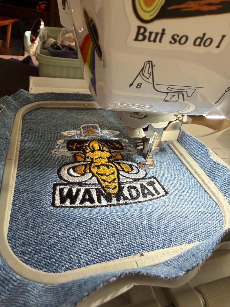 Op de afbeelding: Een witte borduurmachine borduurt een geel en grijs mammoetontwerp met de tekst WANKDAT op een stuk blauwe spijkerstof in een rechthoekige borduurring. De machine is voorzien van een sticker met de tekst But so do I.