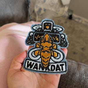 Op de afbeelding: Een geborduurde patch met een centraal goudkleurig bij-achtig wezen met grote vleugels, geplaatst onder een zwarte rechthoekige vorm met een gele hartslaglijn. De tekst WAN DAT is in zwarte letters op een witte achtergrond geborduurd.