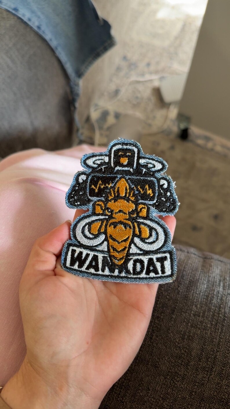 Op de afbeelding: Een geborduurde patch met een centraal goudkleurig bij-achtig wezen met grote vleugels, geplaatst onder een zwarte rechthoekige vorm met een gele hartslaglijn. De tekst WAN DAT is in zwarte letters op een witte achtergrond geborduurd.