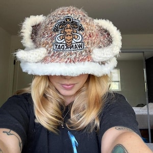 May include: A fuzzy, multi-colored brown and white knit bucket hat featuring a white faux fur trim along the brim and two protruding white plush horns on the sides. A central patch displays a brown mammoth graphic with the text WANKMAAT.