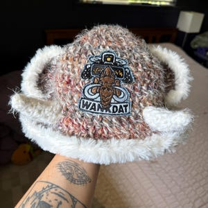 Op de afbeelding: Een bruin en wit gem&ecirc;leerde gebreide muts met een pluizige witte rand en twee uitstekende witte hoorns aan de zijkanten. Een geborduurde patch op de voorkant toont een bruine figuur, een klein geel icoontje en de tekst WAN DAT in zwarte letters.