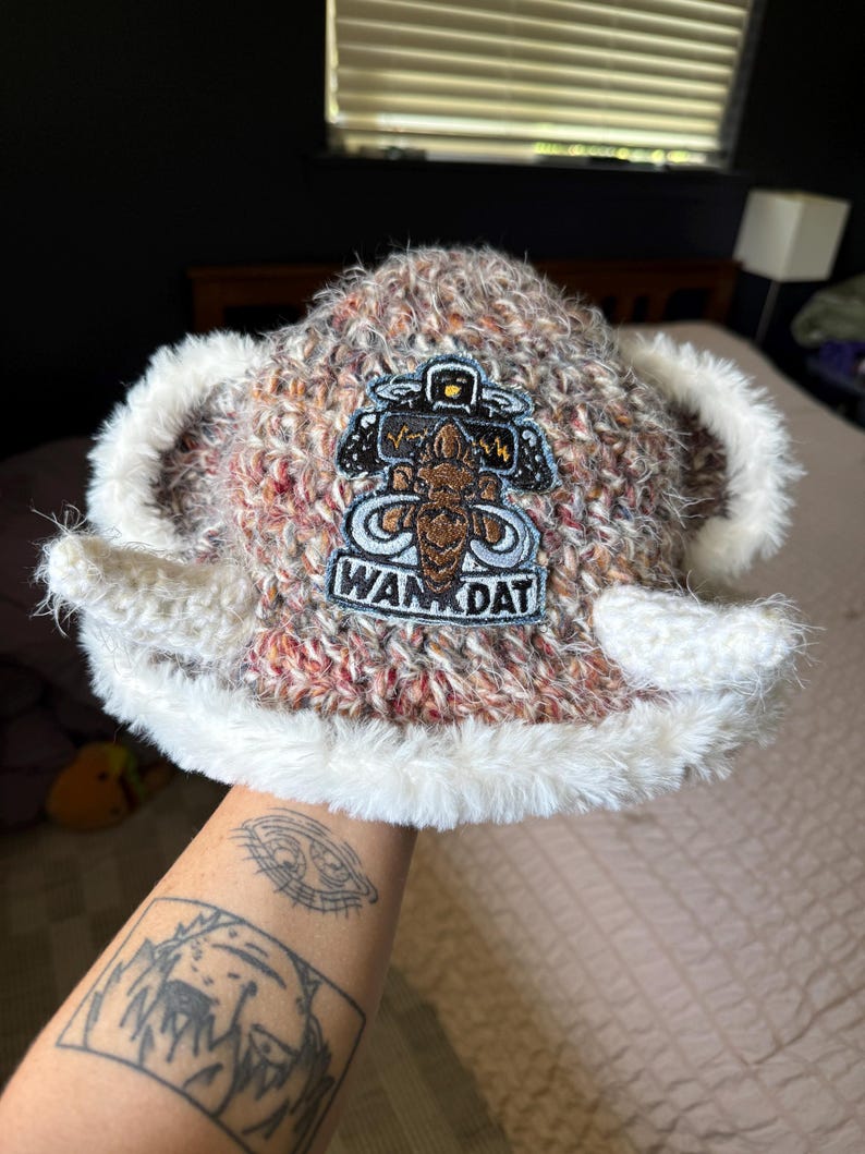 Op de afbeelding: Een bruin en wit gem&ecirc;leerde gebreide muts met een pluizige witte rand en twee uitstekende witte hoorns aan de zijkanten. Een geborduurde patch op de voorkant toont een bruine figuur, een klein geel icoontje en de tekst WAN DAT in zwarte letters.