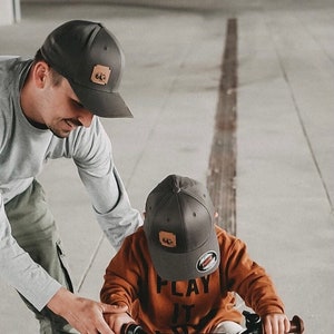 Könnte beinhalten: Ein Kind mit einer dunkelgrauen Baseballkappe und einem orangefarbenen Langarmshirt mit dem Aufdruck "PLAY IT" fährt Fahrrad. Das Kind trägt weiße Turnschuhe. Ein Erwachsener hilft dem Kind. Das Fahrrad hat einen schwarzen Rahmen.