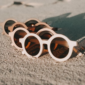 May include: A row of five white round sunglasses with brown lenses, lying on a sandy surface.