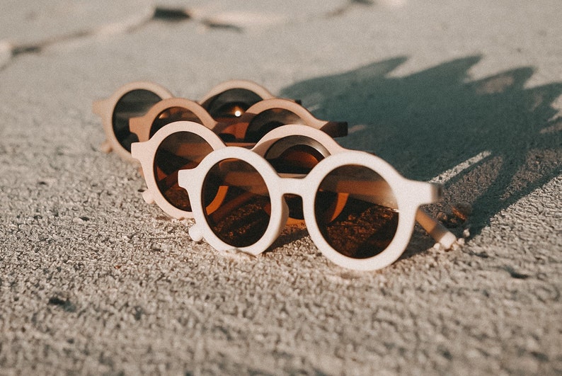 May include: A row of five white round sunglasses with brown lenses, lying on a sandy surface.