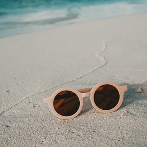 May include: Round pink sunglasses with brown lenses on a white sandy beach.