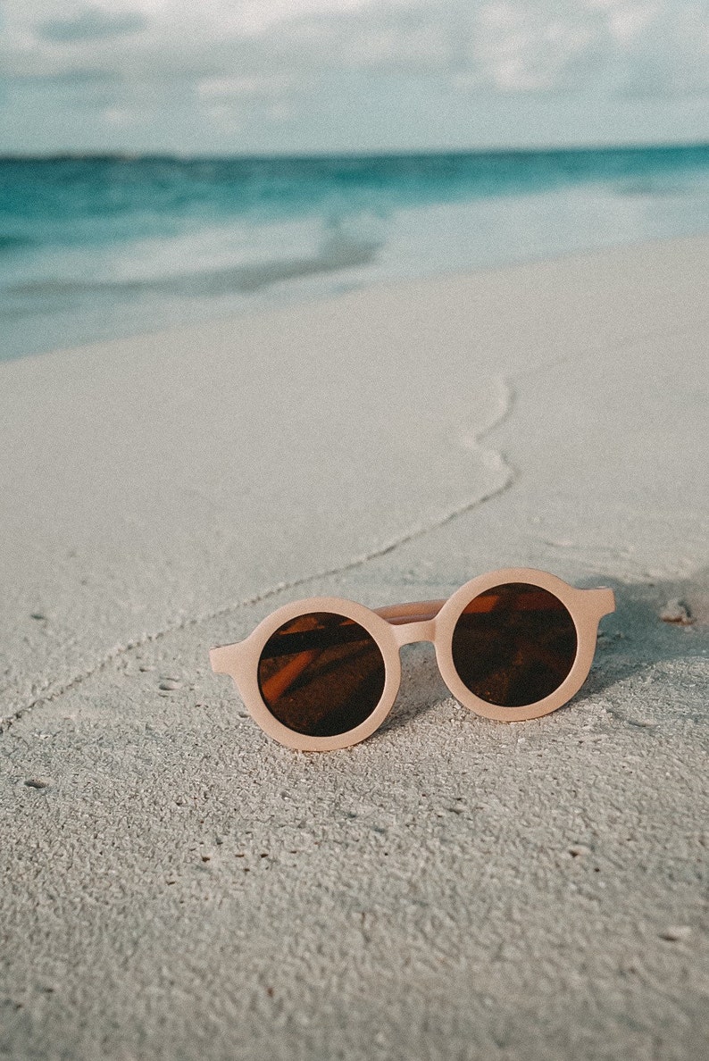 May include: Round pink sunglasses with brown lenses on a white sandy beach.