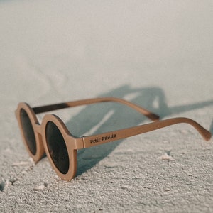 May include: A pair of round, brown sunglasses with dark lenses. The sunglasses are on a white sandy beach. The brand name "Petit Panda" is printed on the side of the sunglasses.