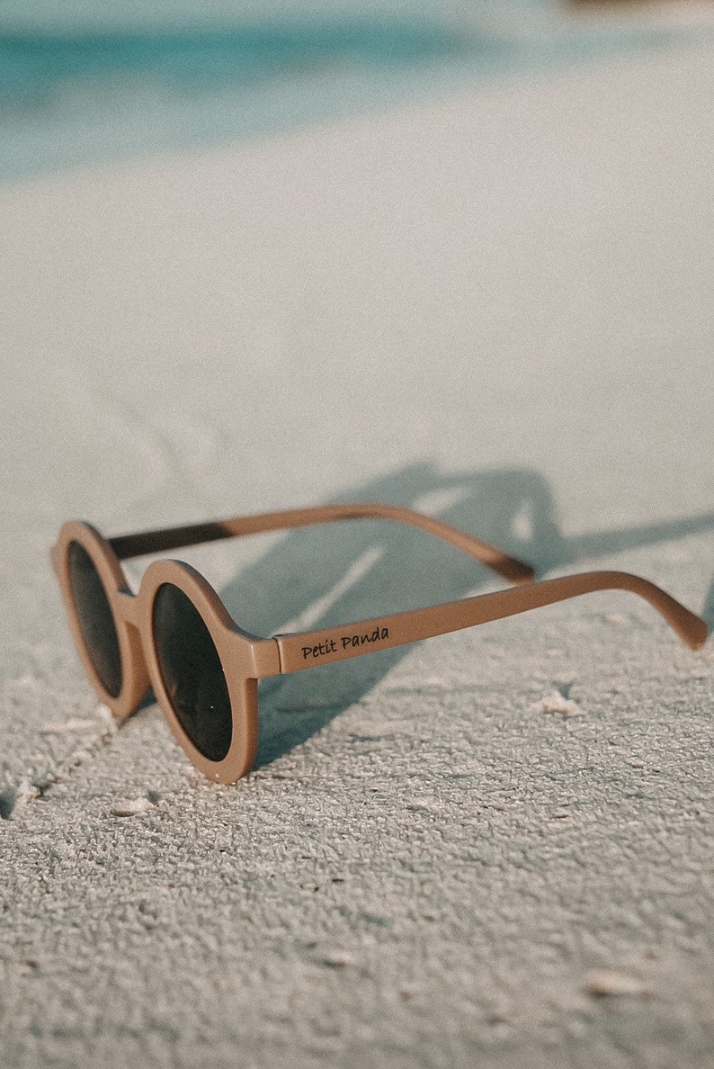 May include: A pair of round, brown sunglasses with dark lenses. The sunglasses are on a white sandy beach. The brand name "Petit Panda" is printed on the side of the sunglasses.