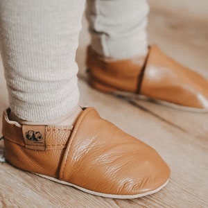 May include: A pair of brown leather baby shoes with a fold-over cuff and a small logo on the side. The shoes are on a wooden floor.