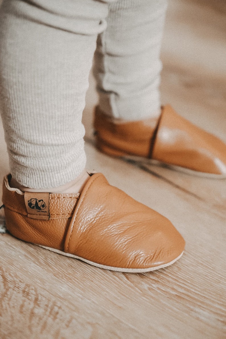 May include: A pair of brown leather baby shoes with a fold-over cuff and a small logo on the side. The shoes are on a wooden floor.