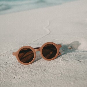 May include: A pair of round brown sunglasses with dark lenses, lying on a white sandy beach.