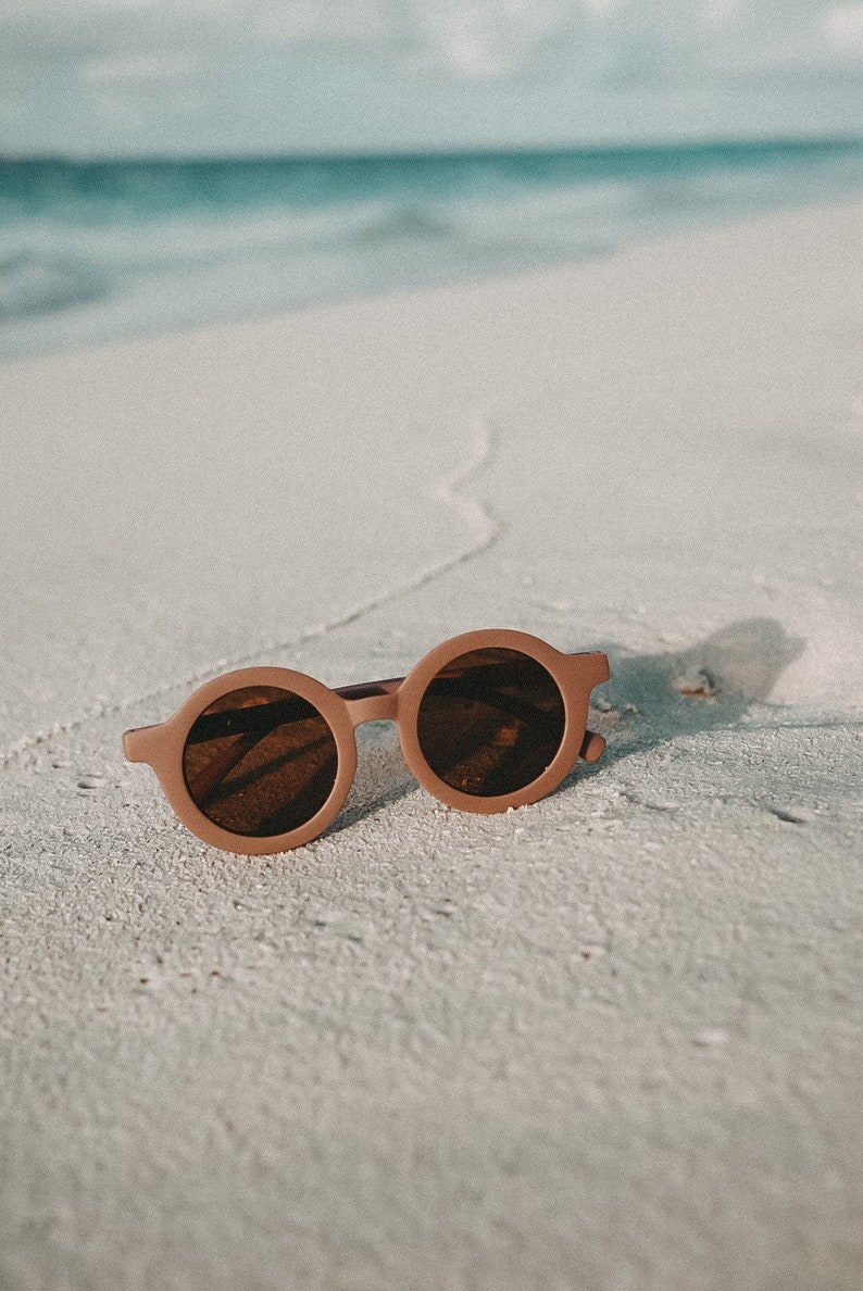 May include: A pair of round brown sunglasses with dark lenses, lying on a white sandy beach.