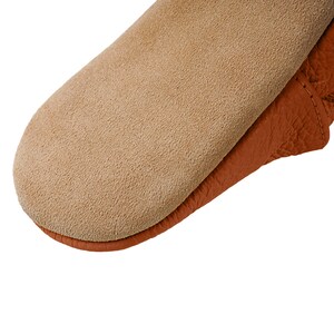 May include: Close-up of a brown leather moccasin with a suede sole. The moccasin has a soft, textured surface and a simple design.
