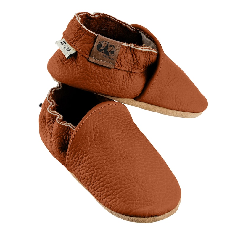 May include: A pair of brown leather baby moccasins with a soft, textured finish. The moccasins have a simple design and are perfect for crawling and walking. The size tag reads "18-24".