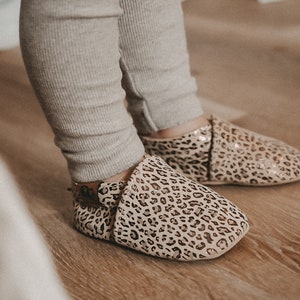 May include: A pair of baby shoes with a leopard print pattern. The shoes are made of a soft, beige material and have a gold glitter finish.