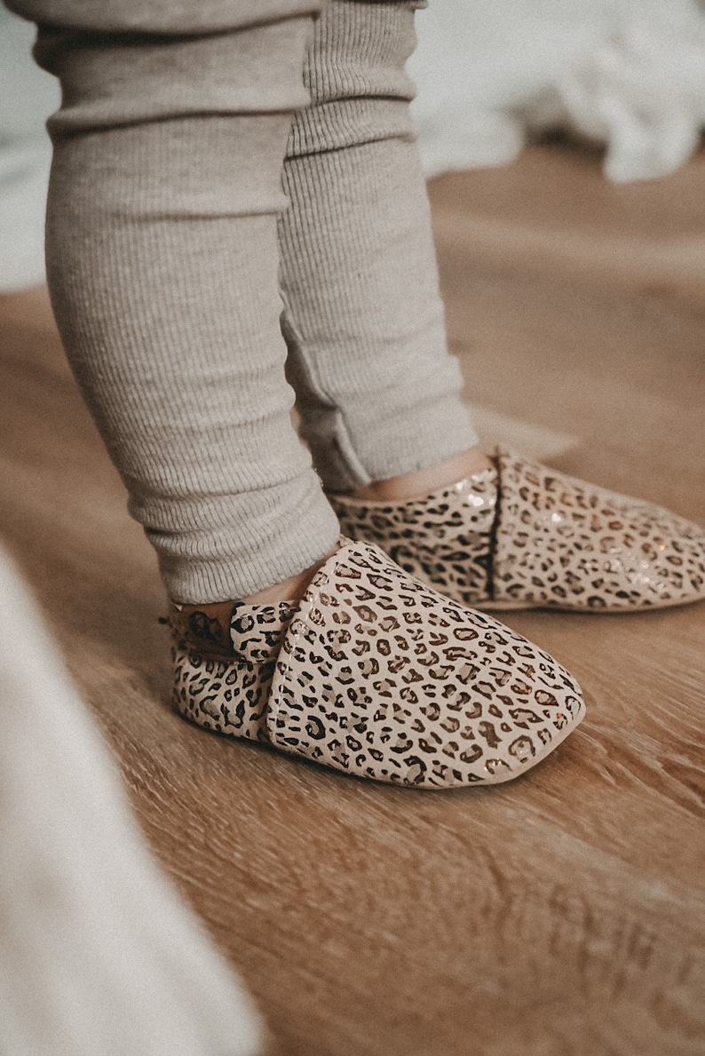 May include: A pair of baby shoes with a leopard print pattern. The shoes are made of a soft, beige material and have a gold glitter finish.