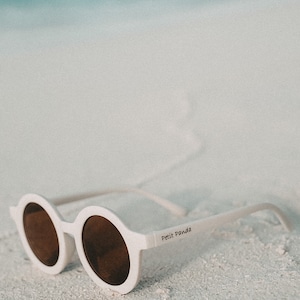 May include: White round sunglasses with brown lenses and the text "Petit Panda" on the side, lying on a white sandy beach.