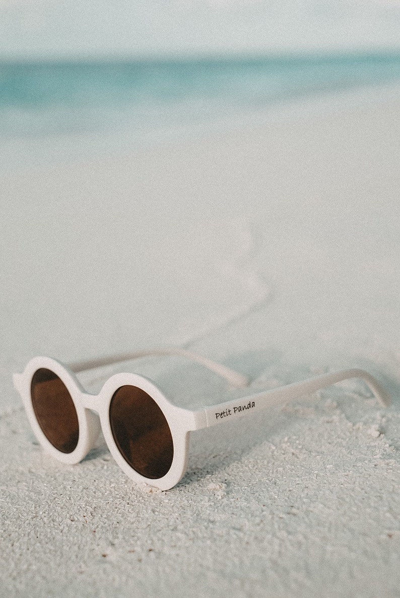 May include: White round sunglasses with brown lenses and the text "Petit Panda" on the side, lying on a white sandy beach.