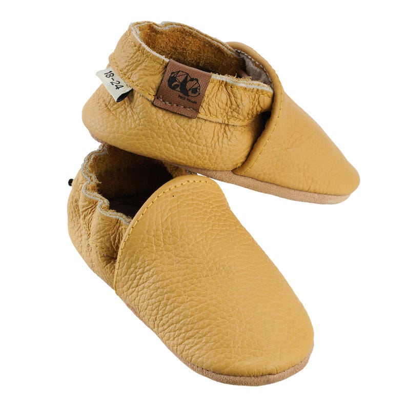 May include: A pair of mustard yellow leather baby shoes with a fold-over top and a size tag that reads "18-24".