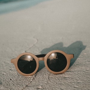 May include: Round brown sunglasses with dark lenses reflecting the sky and water. The sunglasses are lying on a sandy beach.