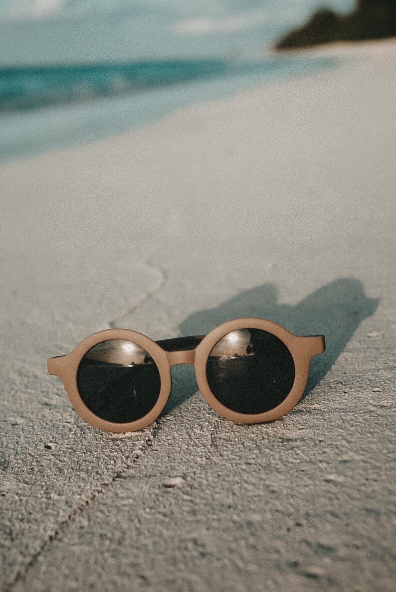 May include: Round brown sunglasses with dark lenses reflecting the sky and water. The sunglasses are lying on a sandy beach.