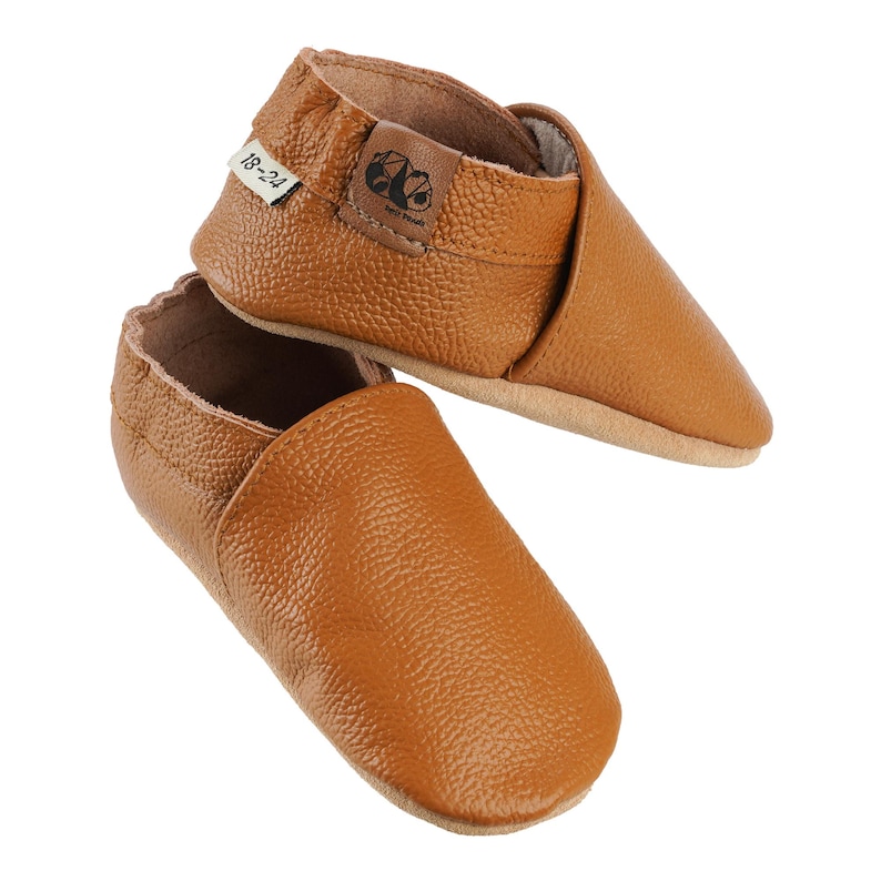 May include: A pair of brown leather baby shoes with a slip-on design. The shoes have a label that says "18-24".