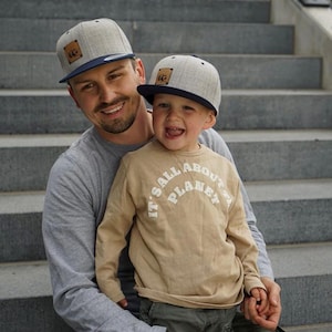 Op de afbeelding: Twee grijze baseballpetten met een marineblauwe klep en een bruin leren patch. Een beige longsleeve met de witte tekst "IT'S ALL ABOUT PLANET". De afbeelding toont een man en een kind.